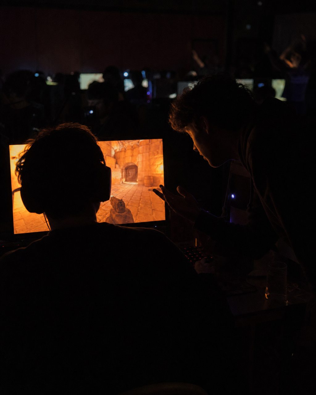 One player leaning over to talk to another at a dark LAN, both lit only by the glowing screens